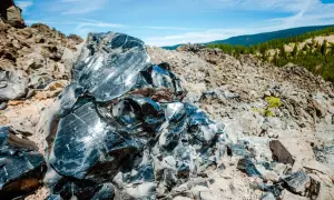 Glänzende Brocken von silber obsidian, die in der Sonne auf einem felsigen Hang glitzern, mit grünen Hügeln im Hintergrund.