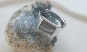 Ein silberner Ring mit einem rechteckigen, grau-weißen Schmuckstein ruht auf einem blauen Kristall.