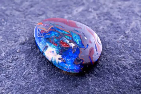 Ein faszinierender Edelstein aus Australien, ein Boulderopal, zeigt ein lebendiges Farbspiel aus Blau, Rot und Orange auf dunklem Grund.