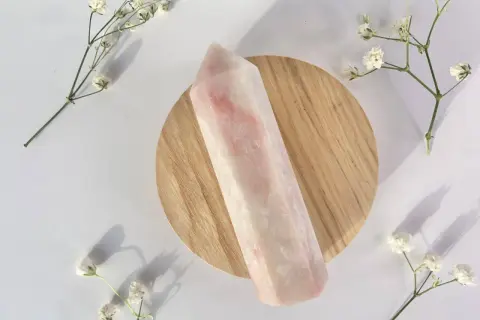 Ein polierter rosa Opal-Kristall liegt auf einem Holzsockel, umgeben von zarten weißen Blüten. Die rosa Opal Wirkung wird oft mit Liebe und Heilung assoziiert.