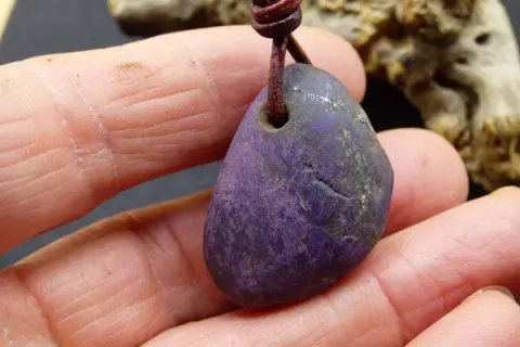 Ein purpurit Anhänger, gehalten in Fingern. Seine tiefviolette Farbe und die natürliche Textur lassen die purpurit wirkung erahnen.