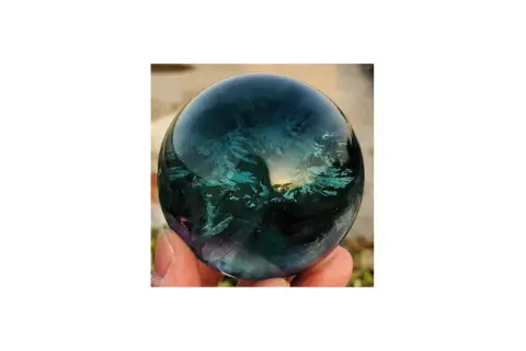Hand hält eine polierte Kugel aus Regenbogenfluorit, die in tiefen Blau- und Grüntönen schimmert. Perfekt, um Regenbogenfluorit kaufen.