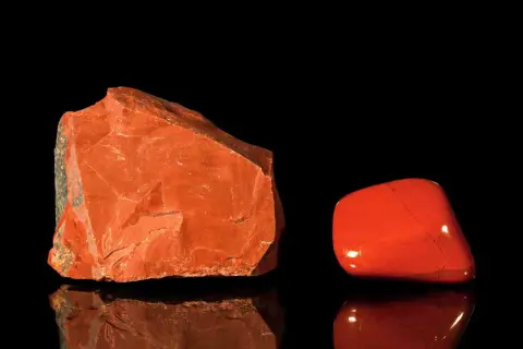 Zwei rote Jaspis-Arten: ein roher Stein und ein polierter Trommelstein, die auf schwarzem Hintergrund reflektieren.