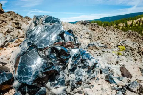 Glänzende Brocken von silber obsidian, die in der Sonne auf einem felsigen Hang glitzern, mit grünen Hügeln im Hintergrund.