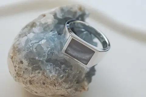 Ein silberner Ring mit einem rechteckigen, grau-weißen Schmuckstein ruht auf einem blauen Kristall.