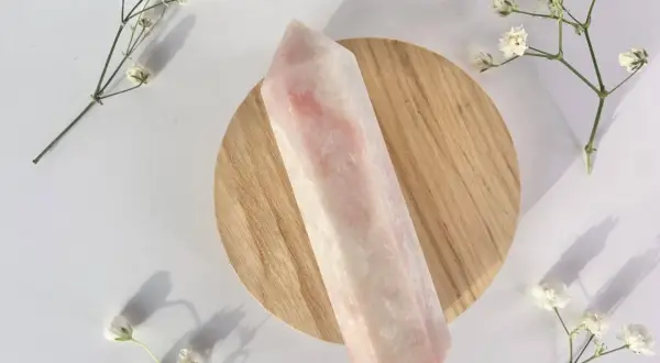 Ein polierter rosa Opal-Kristall liegt auf einem Holzsockel, umgeben von zarten weißen Blüten. Die rosa Opal Wirkung wird oft mit Liebe und Heilung assoziiert.