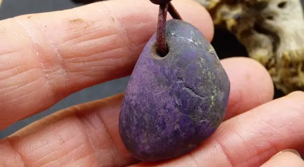 Ein purpurit Anhänger, gehalten in Fingern. Seine tiefviolette Farbe und die natürliche Textur lassen die purpurit wirkung erahnen.