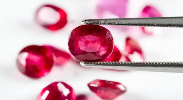 Ein Juwelier hält mit einer Pinzette einen tiefroten Edelstein, der wie der seltenste Diamant der Welt aussieht. Mehrere andere rote Steine liegen unscharf im Hintergrund.