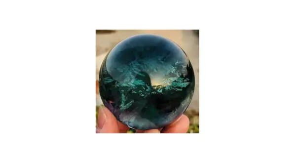 Hand hält eine polierte Kugel aus Regenbogenfluorit, die in tiefen Blau- und Grüntönen schimmert. Perfekt, um Regenbogenfluorit kaufen.