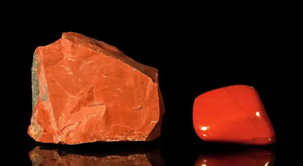Zwei rote Jaspis-Arten: ein roher Stein und ein polierter Trommelstein, die auf schwarzem Hintergrund reflektieren.
