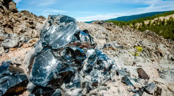 Glänzende Brocken von silber obsidian, die in der Sonne auf einem felsigen Hang glitzern, mit grünen Hügeln im Hintergrund.