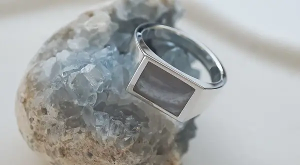 Ein silberner Ring mit einem rechteckigen, grau-weißen Schmuckstein ruht auf einem blauen Kristall.