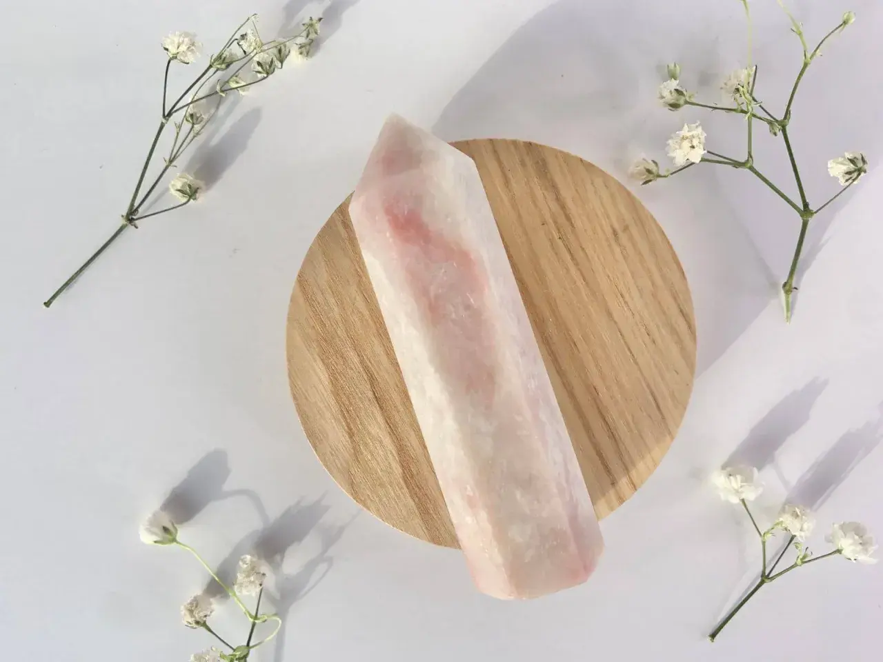 Ein polierter rosa Opal-Kristall liegt auf einem Holzsockel, umgeben von zarten weißen Blüten. Die rosa Opal Wirkung wird oft mit Liebe und Heilung assoziiert.
