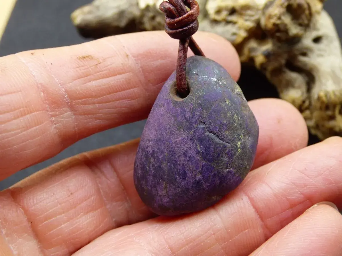 Ein purpurit Anhänger, gehalten in Fingern. Seine tiefviolette Farbe und die natürliche Textur lassen die purpurit wirkung erahnen.