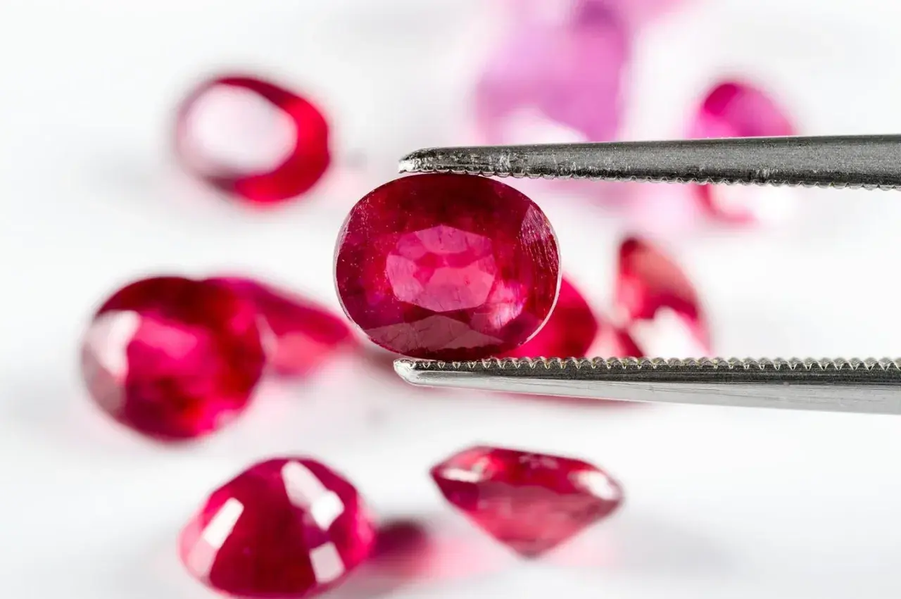 Ein Juwelier hält mit einer Pinzette einen tiefroten Edelstein, der wie der seltenste Diamant der Welt aussieht. Mehrere andere rote Steine liegen unscharf im Hintergrund.