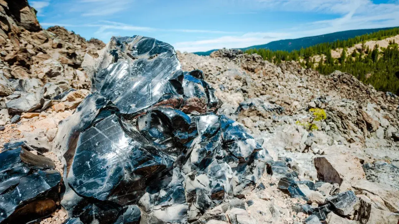 Glänzende Brocken von silber obsidian, die in der Sonne auf einem felsigen Hang glitzern, mit grünen Hügeln im Hintergrund.