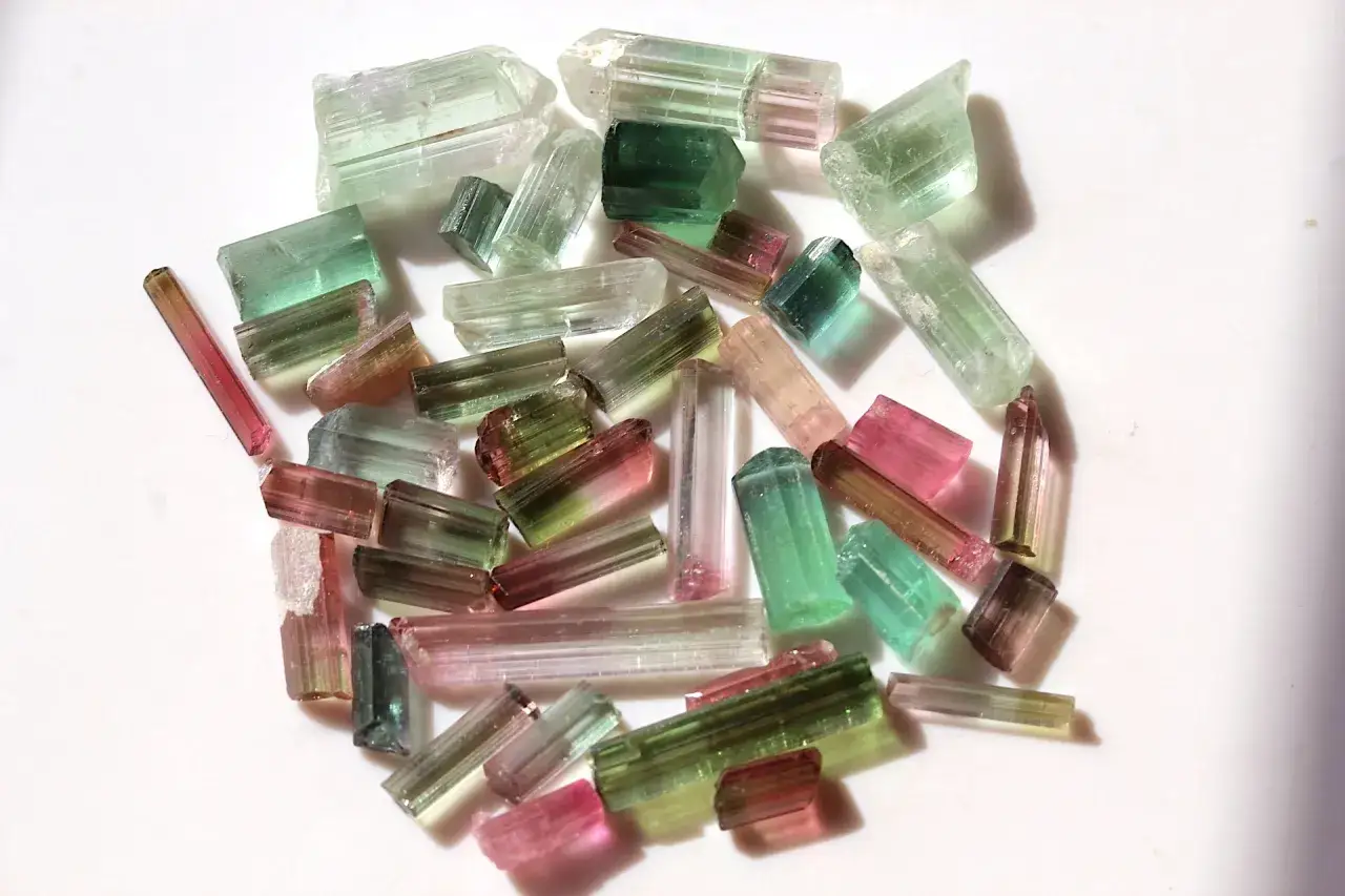 Bunte Turmalin Steine in verschiedenen Grüntönen, Rosa und Transparent liegen auf weißem Untergrund.