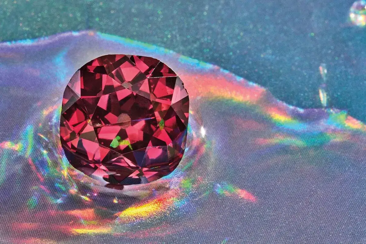 Ein tiefroter, facettierter Edelstein, der wie der seltenste Diamant der Welt schimmert, reflektiert Regenbogenlicht auf schimmerndem Stoff.