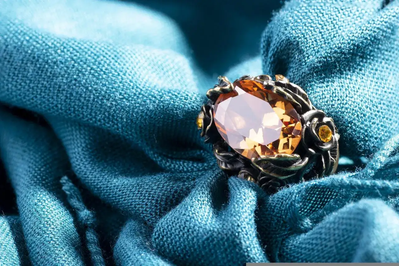 Ein Ring mit einem gro&szlig;en, ovalen, bernsteinfarbenen Stein und kleineren gelben Steinen, eingebettet in ein dunkles Metallgeh&auml;use, liegt auf einem t&uuml;rkisfarbenen Stoff.