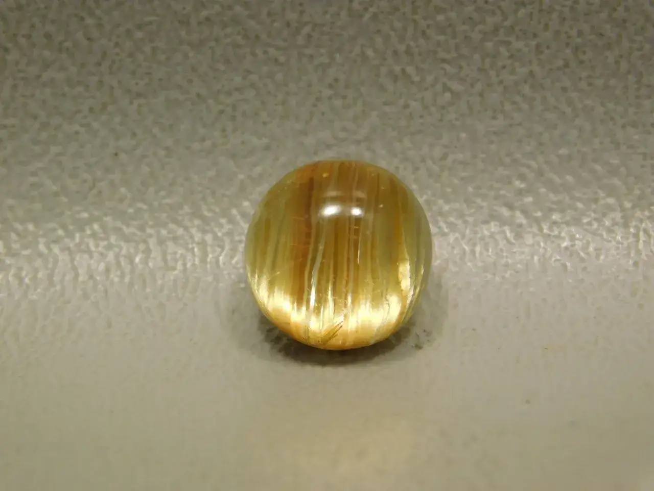 Ein runder, goldener Quarz mit Katzenauge-Effekt schimmert auf hellem Untergrund.