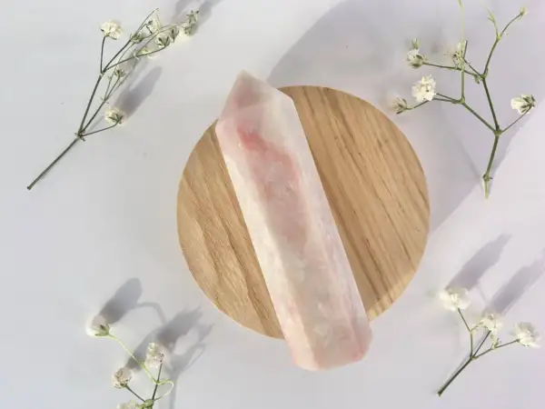 Ein polierter rosa Opal-Kristall liegt auf einem Holzsockel, umgeben von zarten weißen Blüten. Die rosa Opal Wirkung wird oft mit Liebe und Heilung assoziiert.