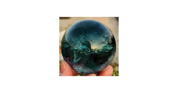 Hand hält eine polierte Kugel aus Regenbogenfluorit, die in tiefen Blau- und Grüntönen schimmert. Perfekt, um Regenbogenfluorit kaufen.