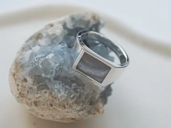 Ein silberner Ring mit einem rechteckigen, grau-weißen Schmuckstein ruht auf einem blauen Kristall.